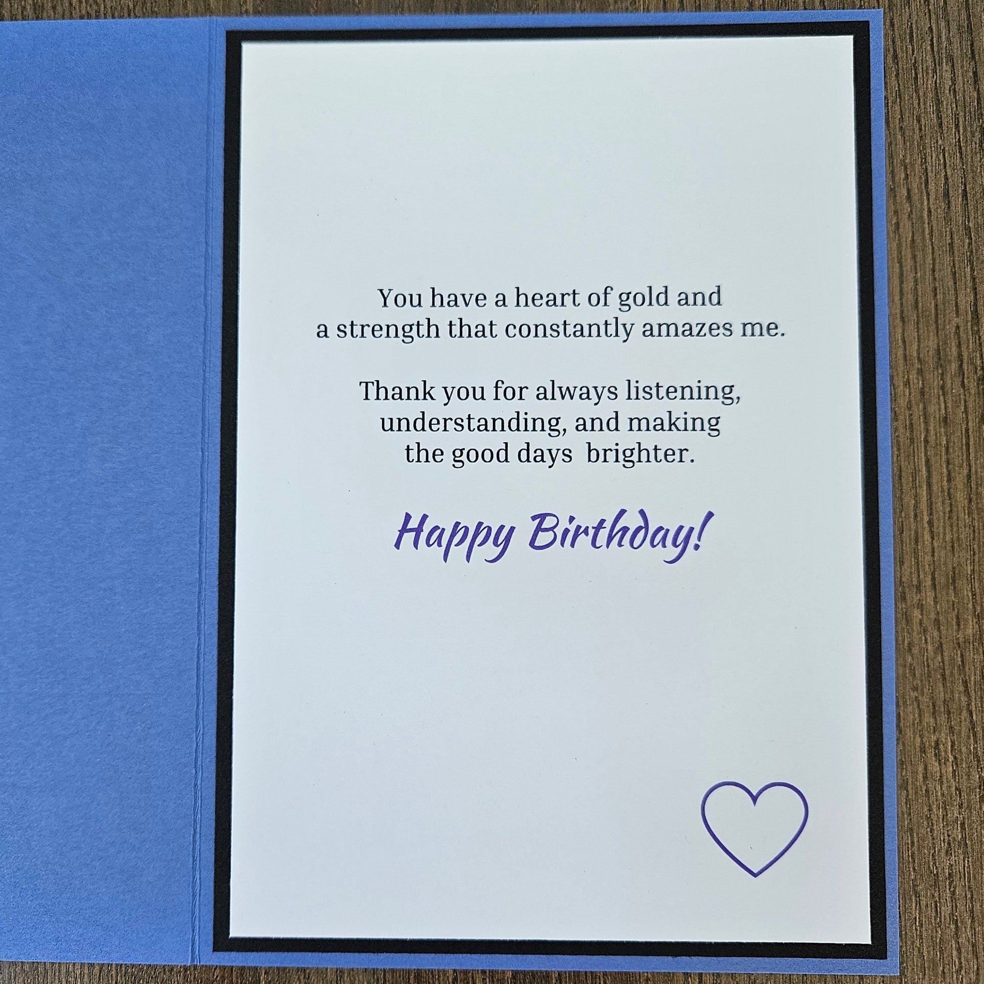 An open square birthday card with a blue outer layer and a white inner card featuring a printed message in black and purple text, plus a small purple heart outline, photographed on a wood surface.