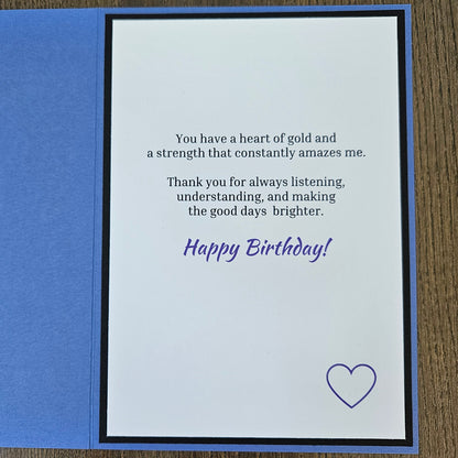 An open square birthday card with a blue outer layer and a white inner card featuring a printed message in black and purple text, plus a small purple heart outline, photographed on a wood surface.