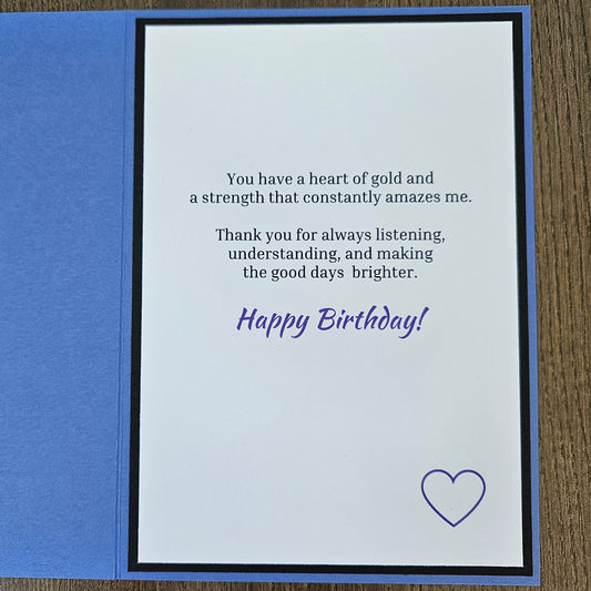 An open square birthday card with a blue outer layer and a white inner card featuring a printed message in black and purple text, plus a small purple heart outline, photographed on a wood surface.