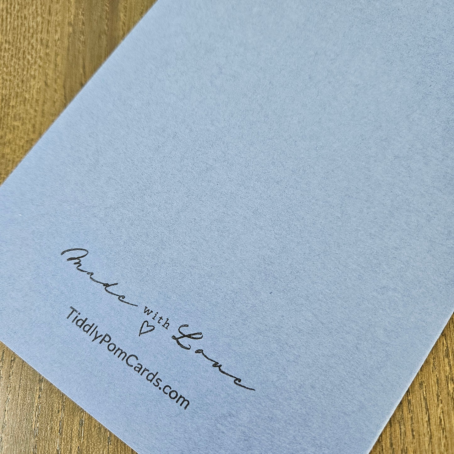 A close-up of a light blue textured paper card printed with the words 'Proud with Love' and 'TiddlyPomCards.com' in black script, resting on a wooden surface.