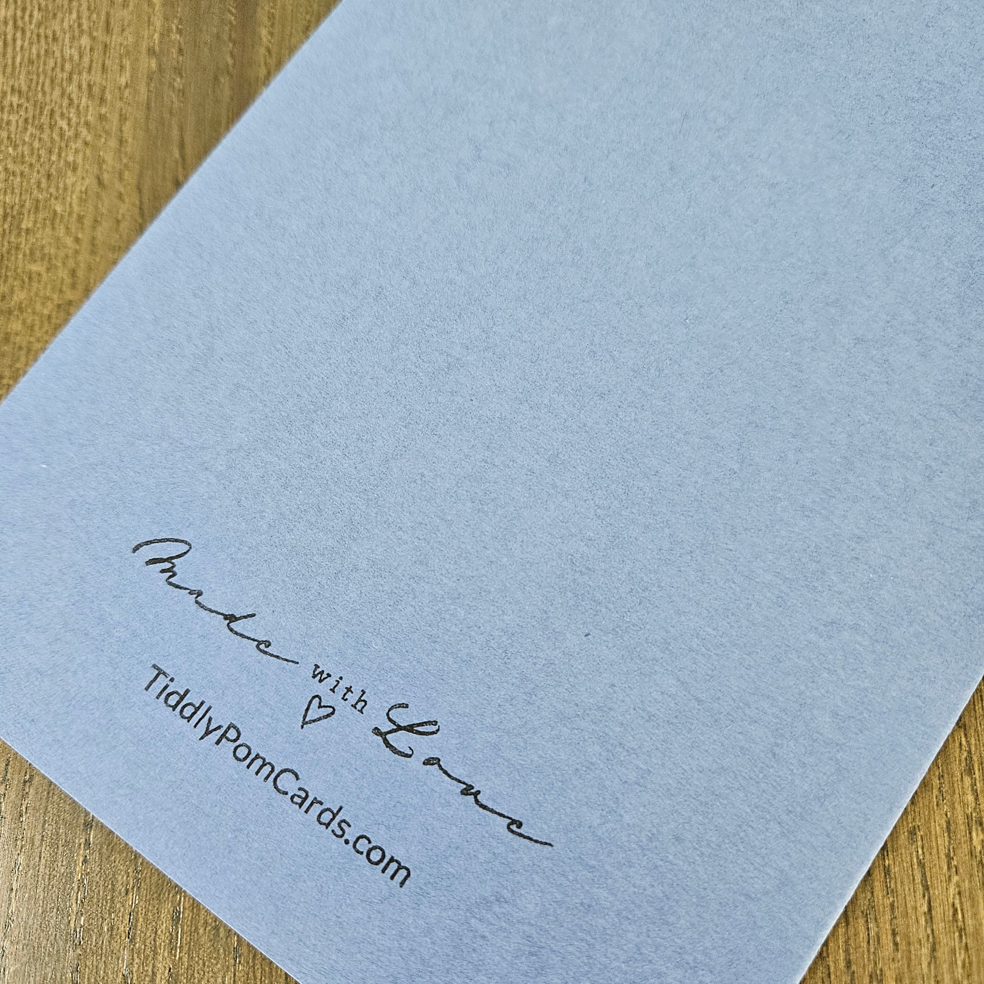 A close-up of a light blue textured paper card printed with the words 'Proud with Love' and 'TiddlyPomCards.com' in black script, resting on a wooden surface.
