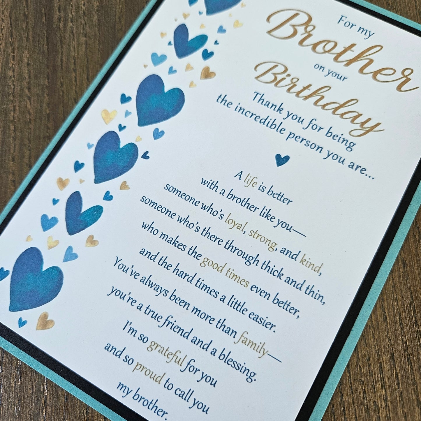 A close-up of a printed birthday card or gift message card laid on a wooden surface. The card has a white background with blue and gold heart illustrations, blue and gold script text reading 'For my Brother on your Birthday' and a sentimental message for a brother.