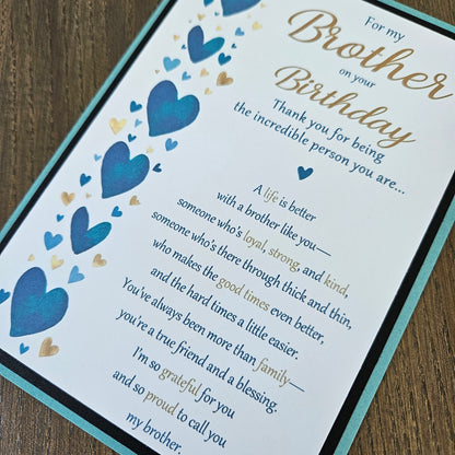 A close-up of a printed birthday card or gift message card laid on a wooden surface. The card has a white background with blue and gold heart illustrations, blue and gold script text reading 'For my Brother on your Birthday' and a sentimental message for a brother.