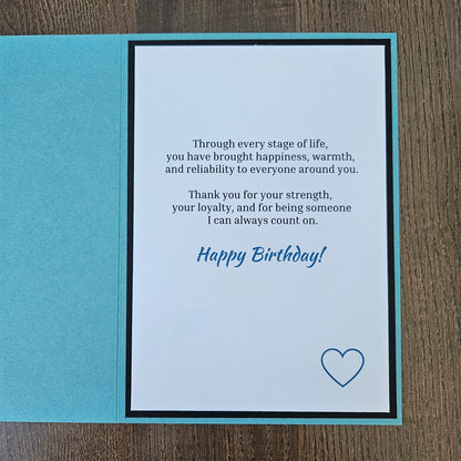 A square folded greeting card shown open on a wood surface, with a turquoise outer panel and a white inner panel printed with a birthday message in black and blue text plus a small blue heart outline.