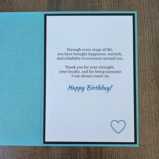 A square folded greeting card shown open on a wood surface, with a turquoise outer panel and a white inner panel printed with a birthday message in black and blue text plus a small blue heart outline.