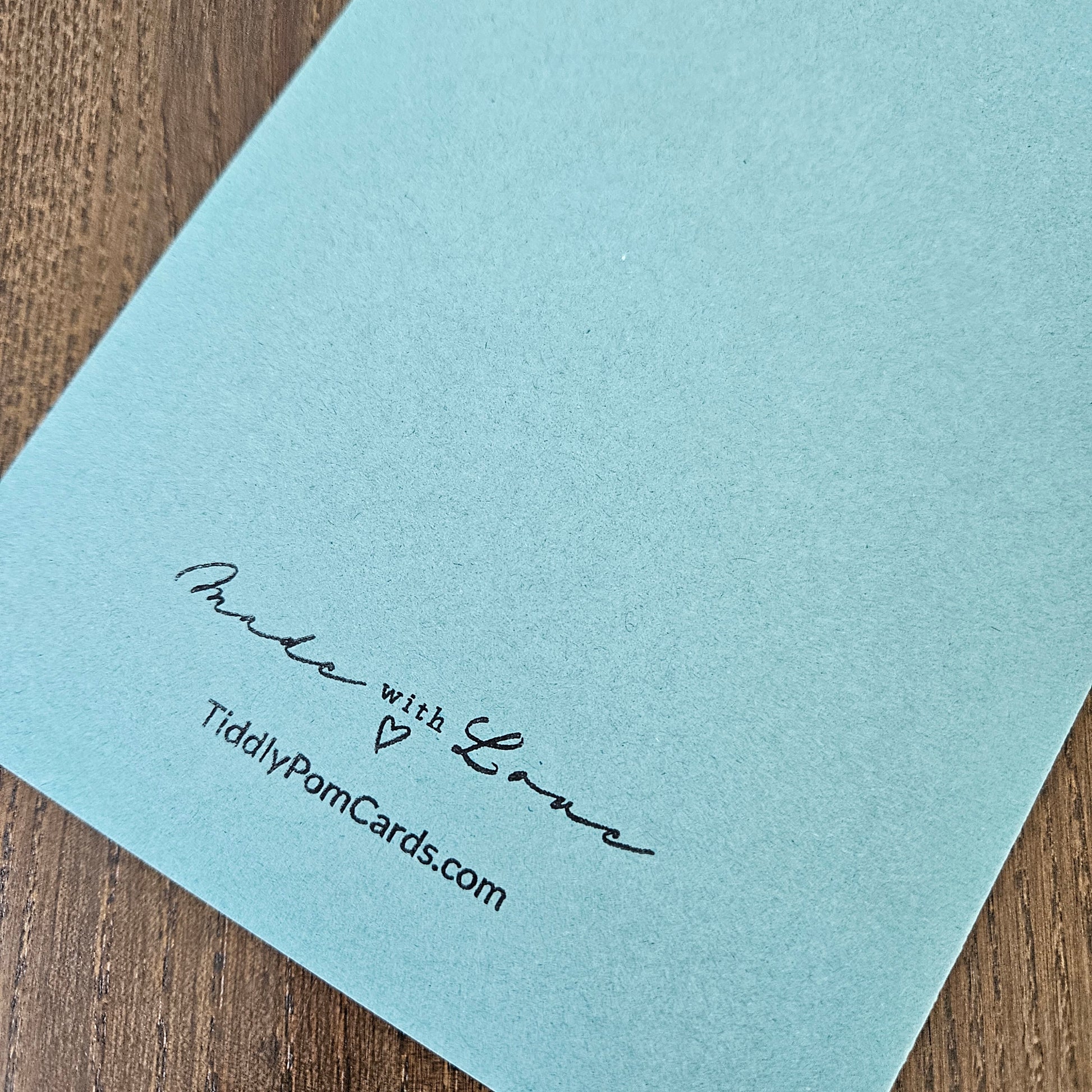 A close-up of a light blue card or envelope corner printed with the words 'Muda with Love' and 'TiddlyPomCards.com' in black script, placed on a brown wood surface.