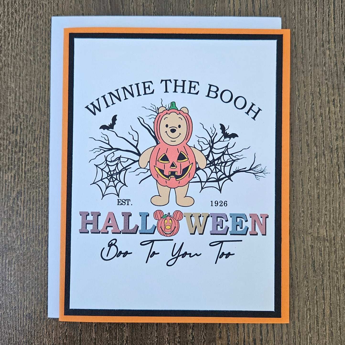A Halloween greeting card featuring Winnie the Boo, a teddy bear in a pumpkin costume, with the text 'WINNIE THE BOOH HALLOWEEN Boo To You Too' on a white card with an orange and black border, placed on a brown wood background.