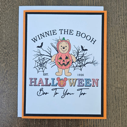 A Halloween greeting card featuring Winnie the Boo, a teddy bear in a pumpkin costume, with the text 'WINNIE THE BOOH HALLOWEEN Boo To You Too' on a white card with an orange and black border, placed on a brown wood background.