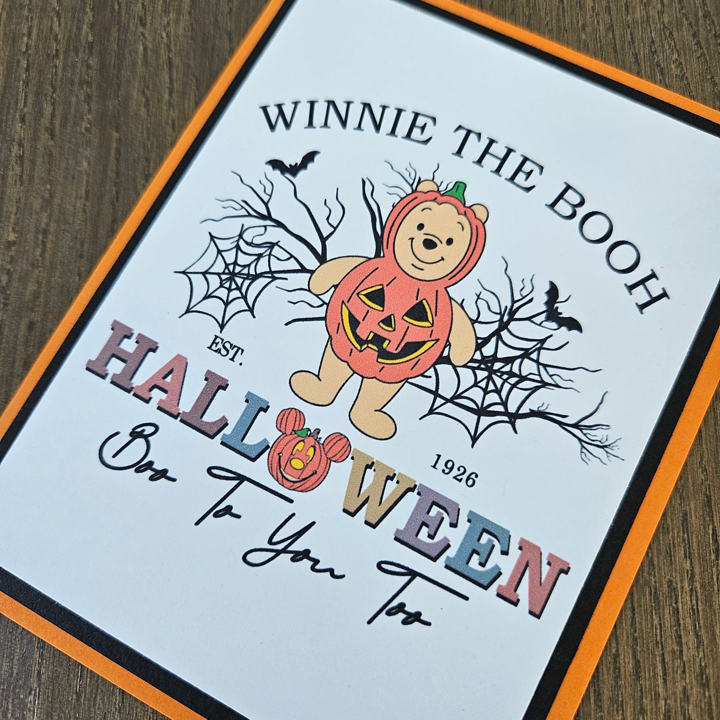 A Halloween greeting card featuring Winnie the Pooh in a pumpkin costume, surrounded by spiderwebs, bats, and the text 'WINNIE THE BOOTH HALLOWEEN Boo To You Too', photographed on a wooden surface.