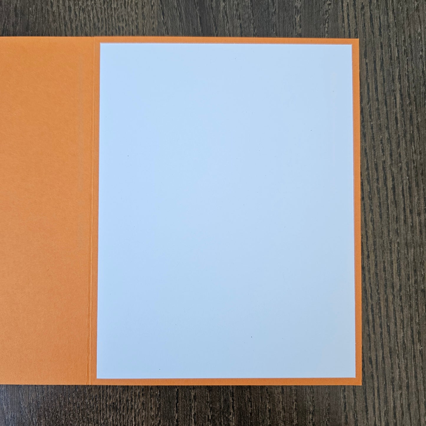 An open orange greeting card with blank white paper inside, placed on a dark wood-grained surface.