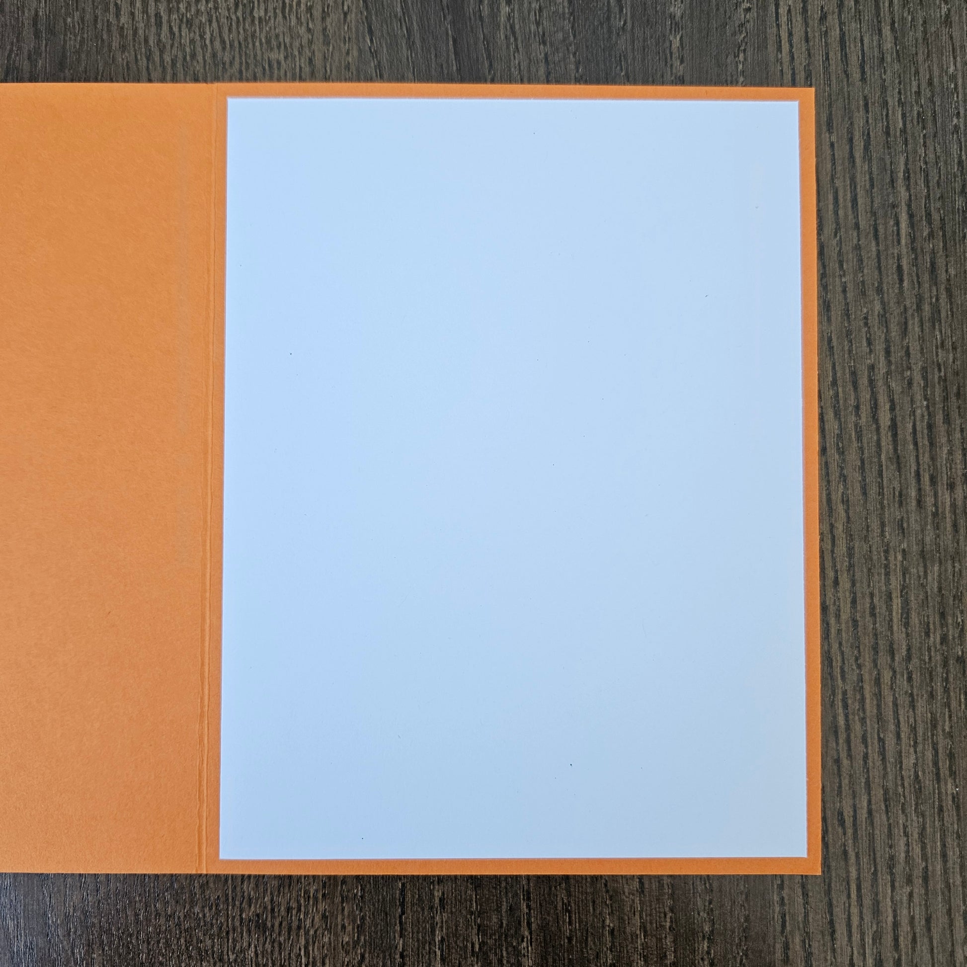 An open orange greeting card with blank white paper inside, placed on a dark wood-grained surface.