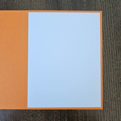 An open orange greeting card with blank white paper inside, placed on a dark wood-grained surface.