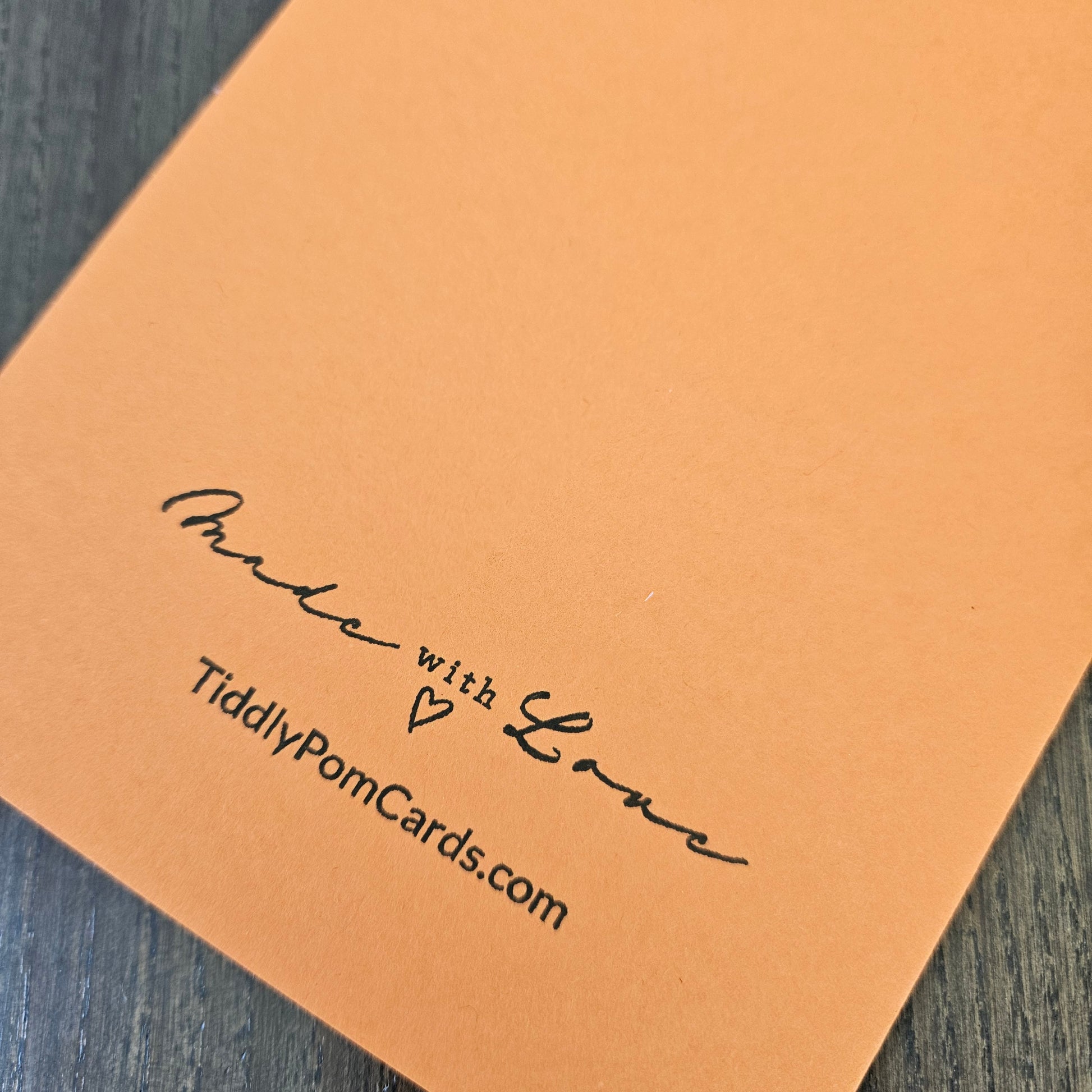 A close-up of a peach-colored greeting card or envelope corner printed with the text 'Made with Love' and 'TiddlyPomCards.com' in black script, resting on a dark wood surface.