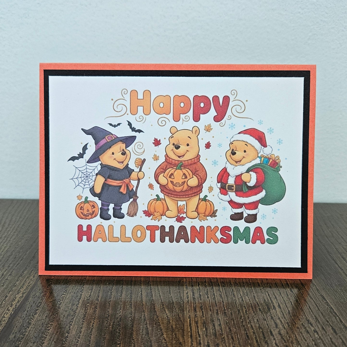 A greeting card with a Halloween and Thanksgiving holiday design, featuring Winnie the Pooh characters in costume, placed on a dark wooden surface against a light gray wall.