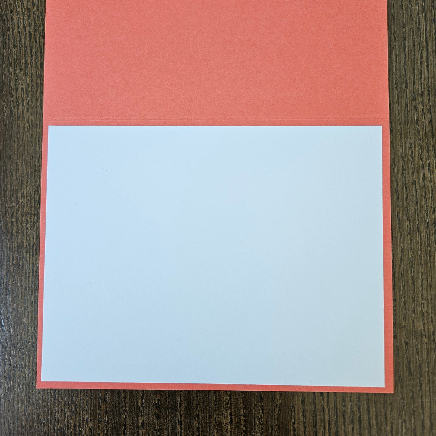 A square greeting card or paper craft template with a coral red border and a large blank white center, displayed flat on a dark wood background.