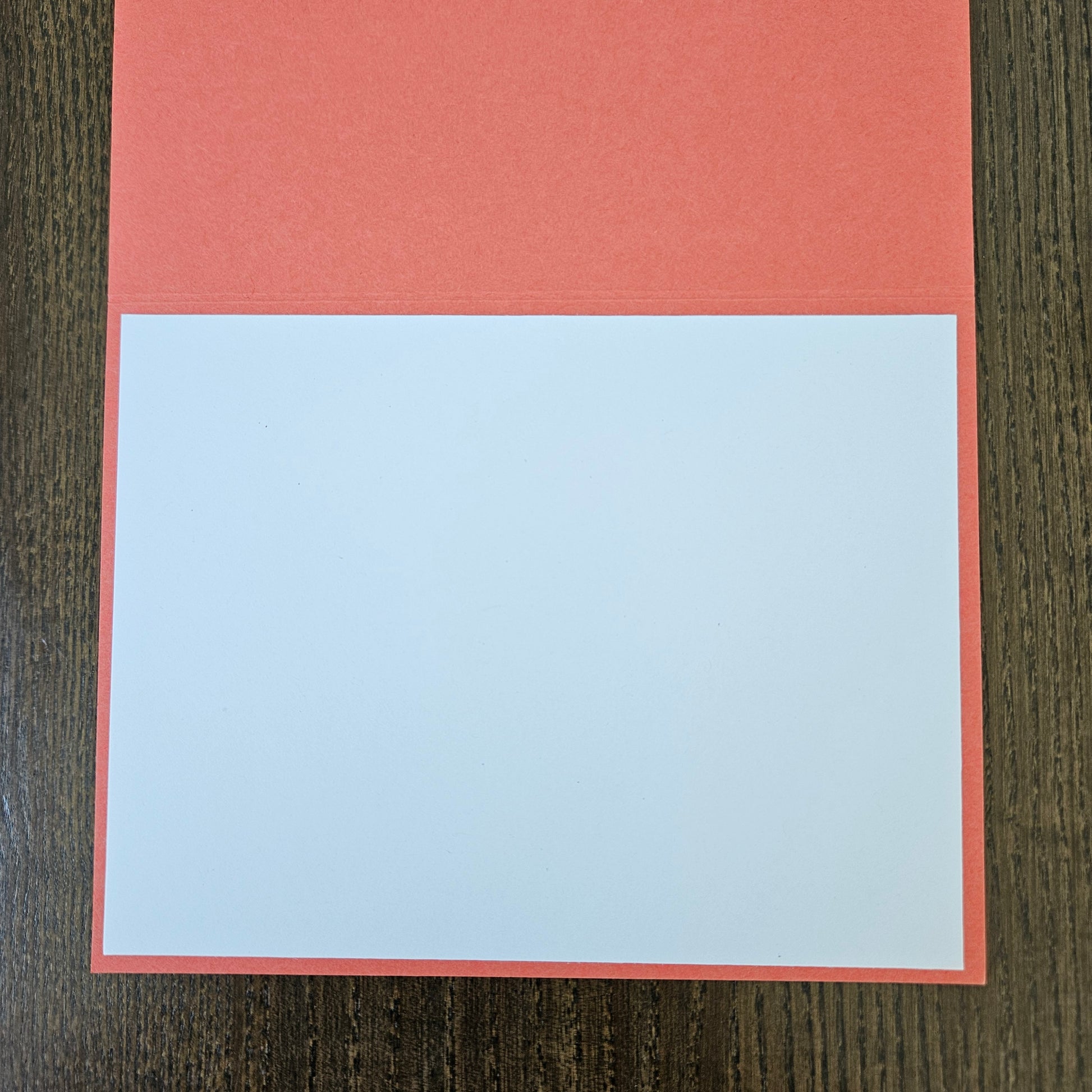 A square greeting card or paper craft template with a coral red border and a large blank white center, displayed flat on a dark wood background.