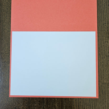 A square greeting card or paper craft template with a coral red border and a large blank white center, displayed flat on a dark wood background.