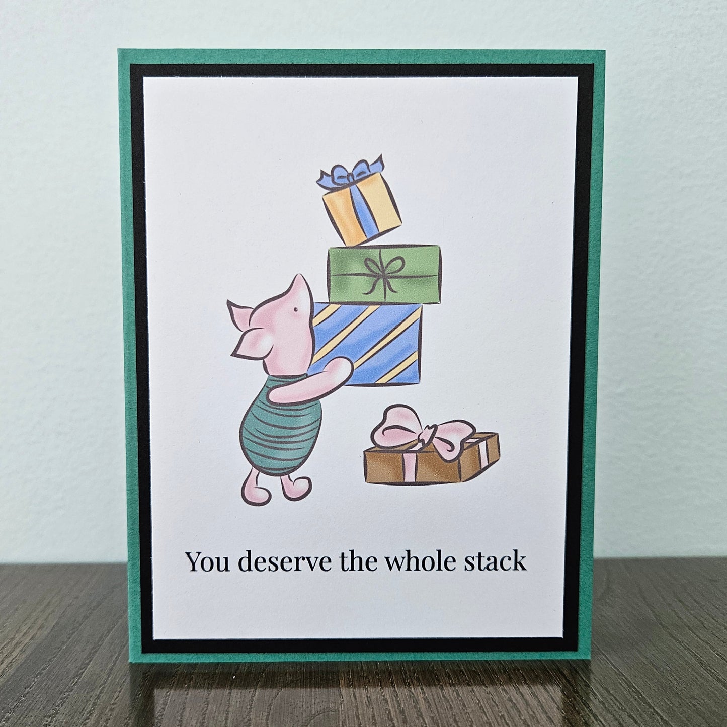 Front of a handmade A2 birthday card featuring classic Piglet balancing a stack of wrapped birthday gifts framed by black and green borders with text reading You deserve the whole stack.