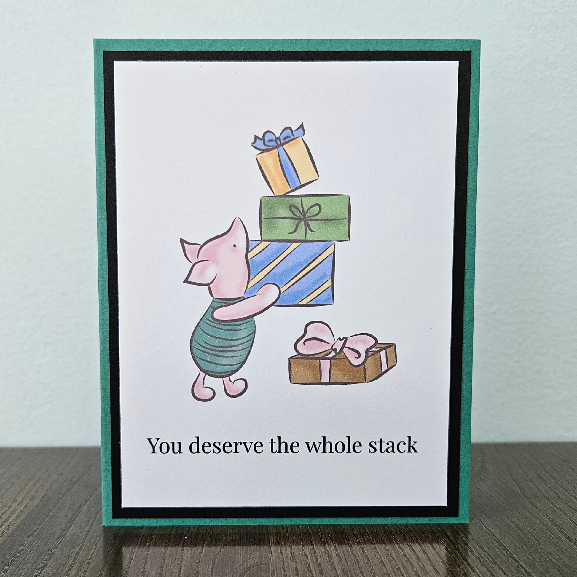 Front of a handmade A2 birthday card featuring classic Piglet balancing a stack of wrapped birthday gifts framed by black and green borders with text reading You deserve the whole stack.