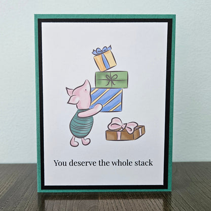Front of a handmade A2 birthday card featuring classic Piglet balancing a stack of wrapped birthday gifts framed by black and green borders with text reading You deserve the whole stack.