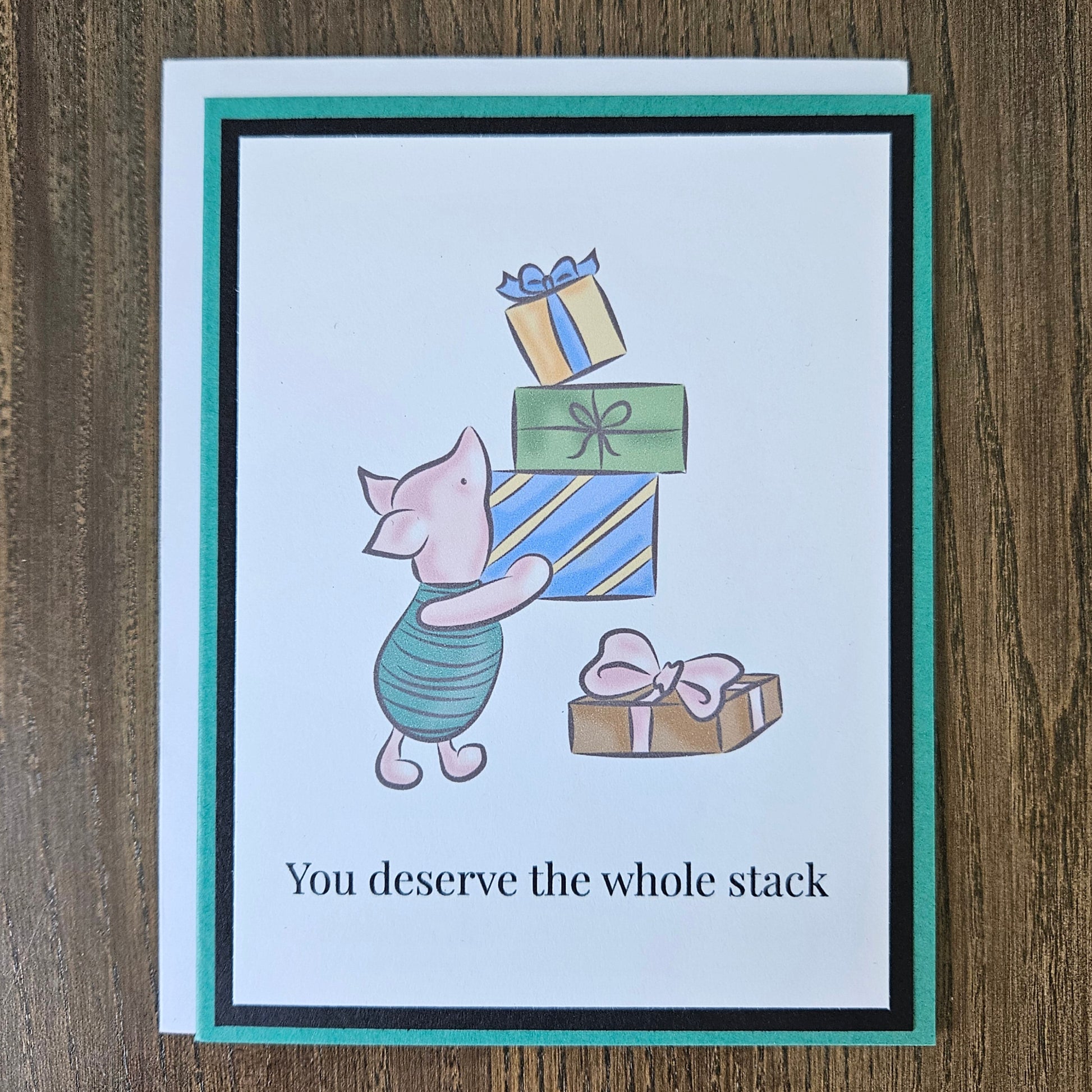 Front of a handmade A2 birthday card featuring classic Piglet balancing a stack of wrapped birthday gifts framed by black and green borders with text reading You deserve the whole stack.
