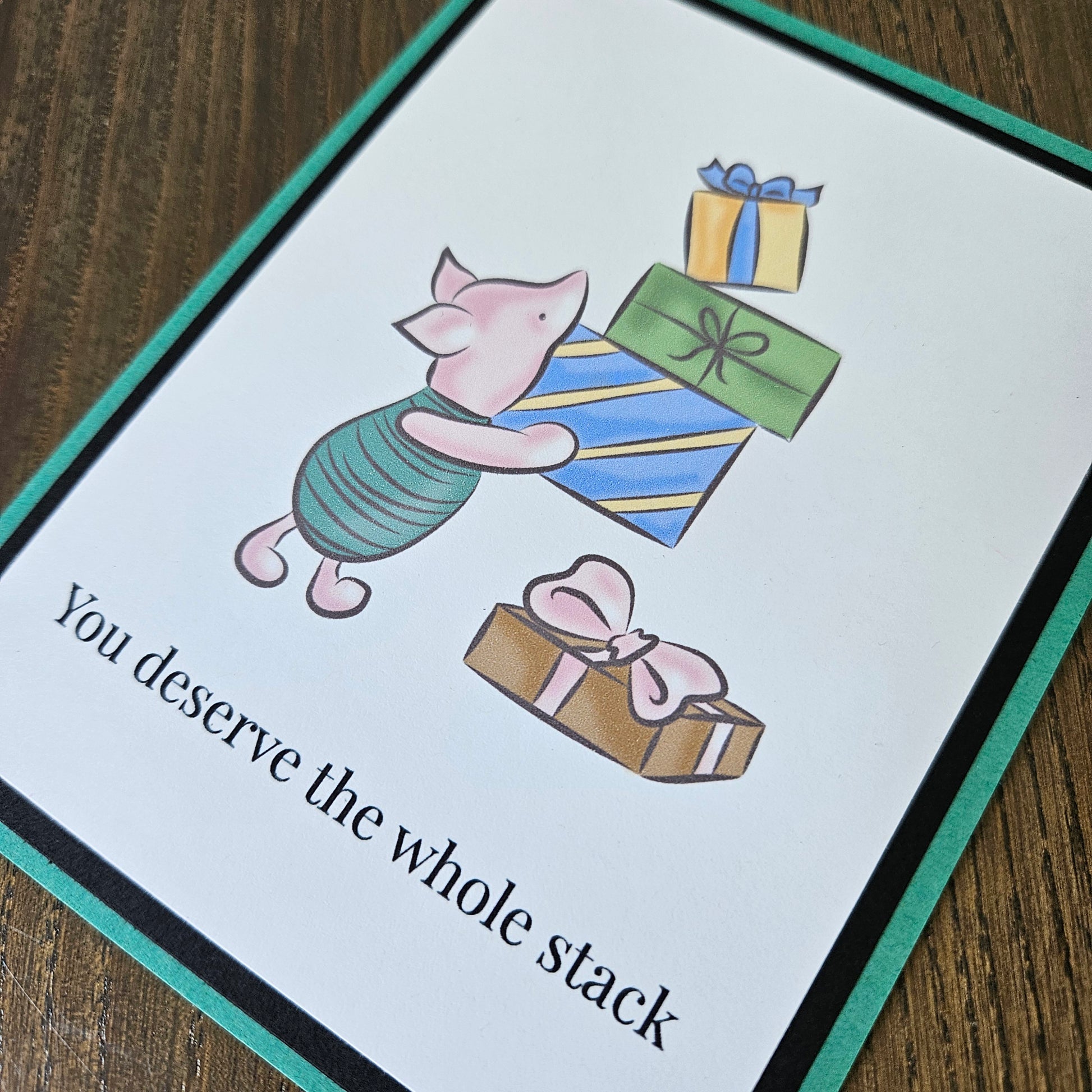 Front of a handmade A2 birthday card featuring classic Piglet balancing a stack of wrapped birthday gifts framed by black and green borders with text reading You deserve the whole stack.