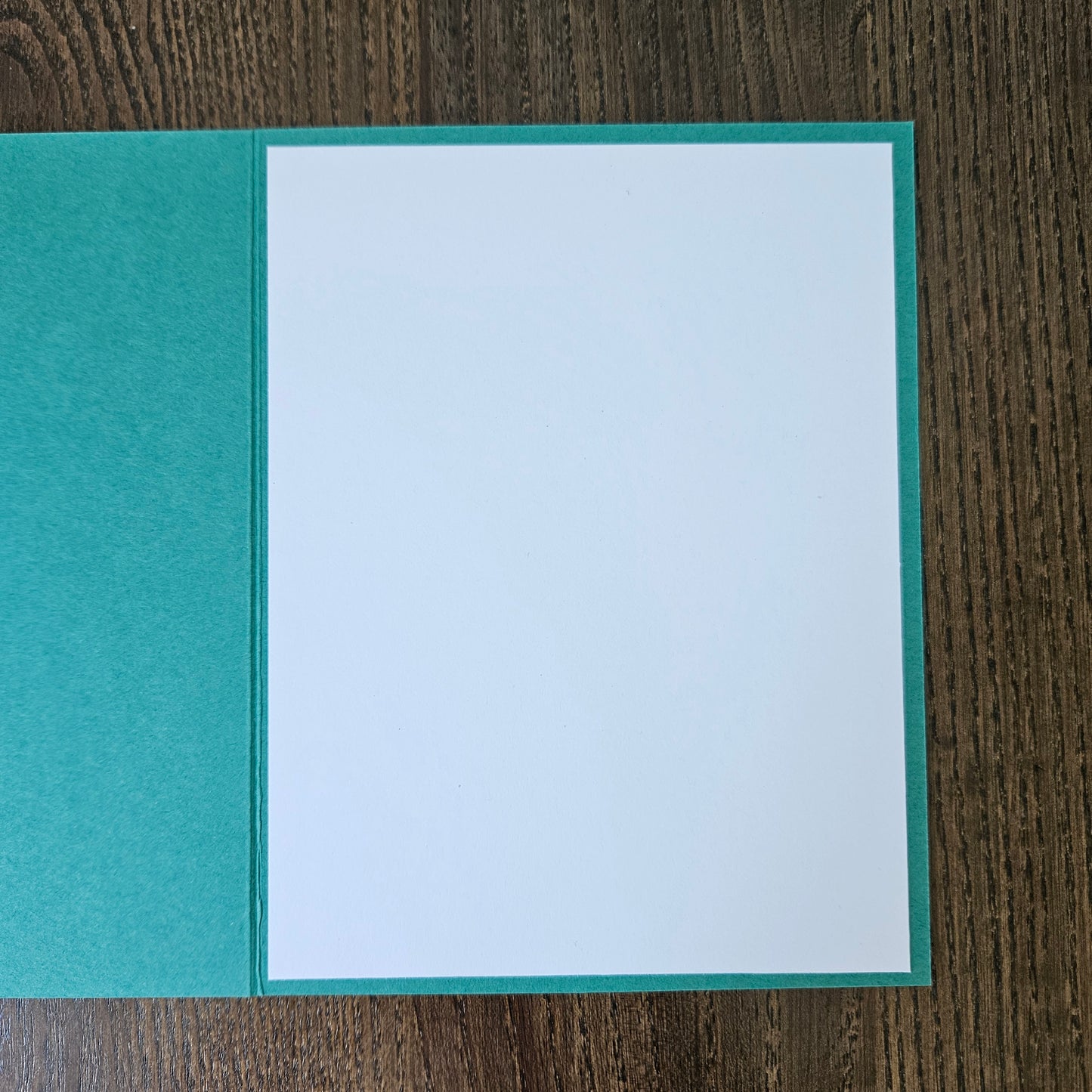 An open greeting card with a textured teal-green cover and a blank white insert page, photographed from above on a dark wood surface.