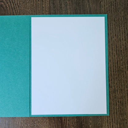 An open greeting card with a textured teal-green cover and a blank white insert page, photographed from above on a dark wood surface.