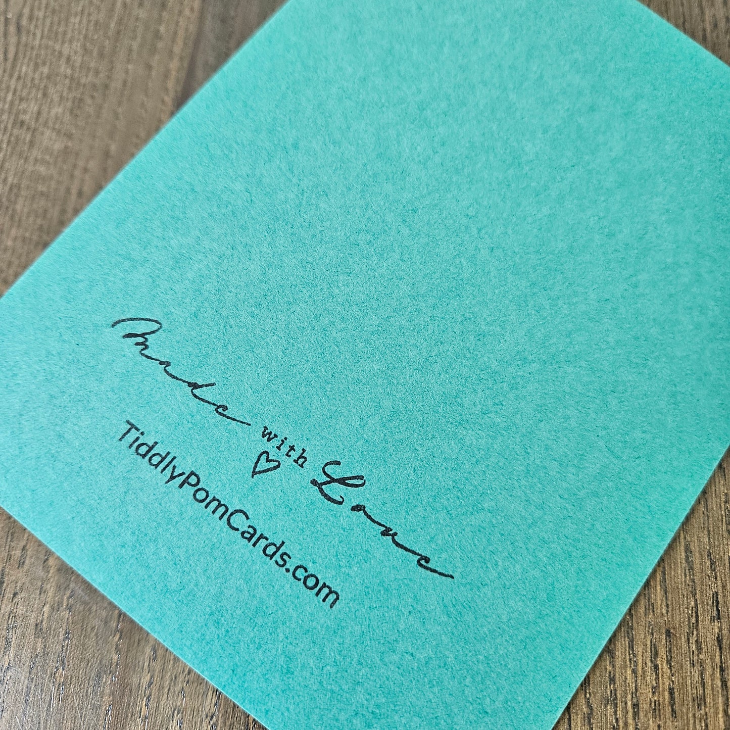 A close-up of a turquoise card on a wooden surface, printed with the text 'Praise with Love' and 'TiddlyPornCards.com' in black script.