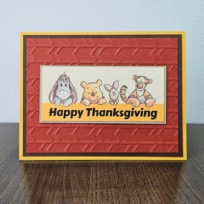 Front of a handmade A2 Thanksgiving card featuring classic Winnie the Pooh, Eeyore, Piglet, and Tigger on an embossed rust-red panel framed by yellow and brown borders with text reading Happy Thanksgiving.