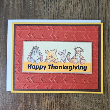 Front of a handmade A2 Thanksgiving card featuring classic Winnie the Pooh, Eeyore, Piglet, and Tigger on an embossed rust-red panel framed by yellow and brown borders with text reading Happy Thanksgiving.