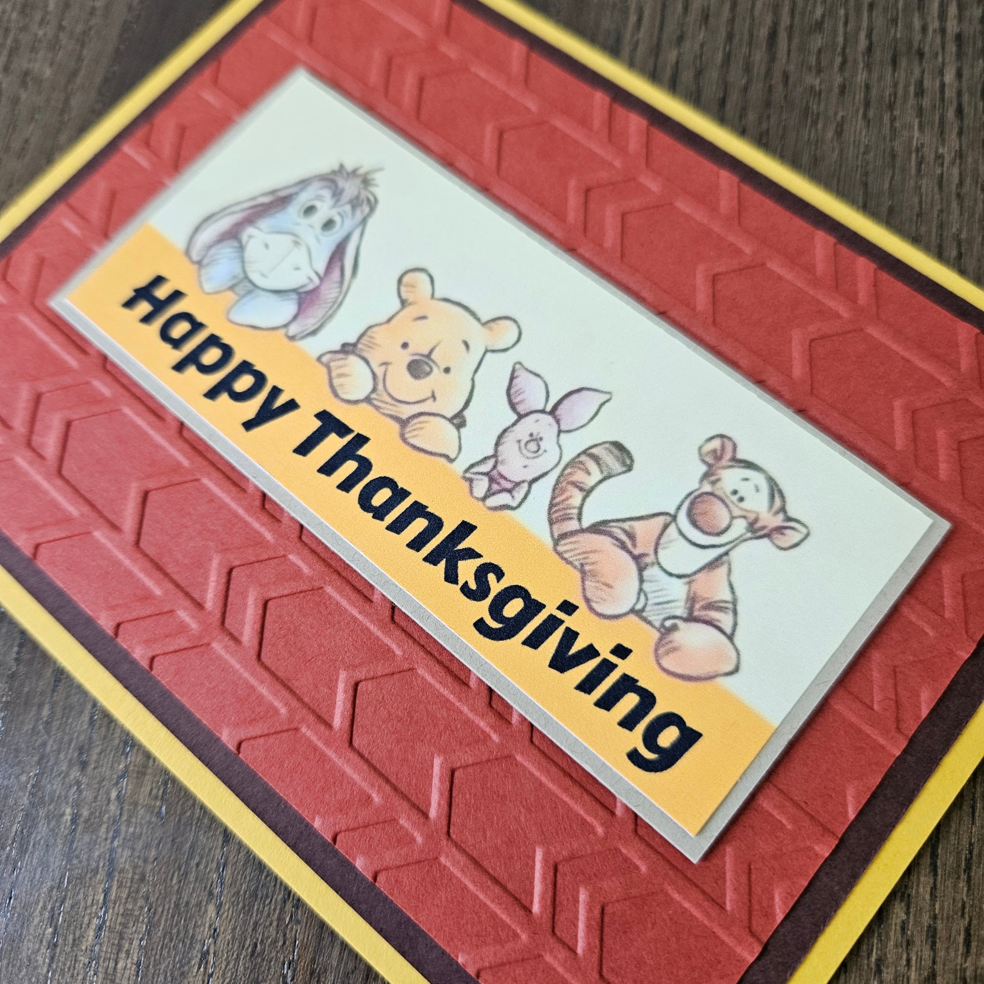 A handmade Thanksgiving greeting card featuring embossed red and brown geometric patterns, a yellow border, and a white banner with cartoon characters including Winnie the Pooh, Tigger, Piglet, and Eeyore, with the text 'Happy Thanksgiving' on a wooden surface.