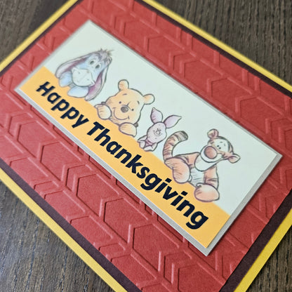 A handmade Thanksgiving greeting card featuring embossed red and brown geometric patterns, a yellow border, and a white banner with cartoon characters including Winnie the Pooh, Tigger, Piglet, and Eeyore, with the text 'Happy Thanksgiving' on a wooden surface.