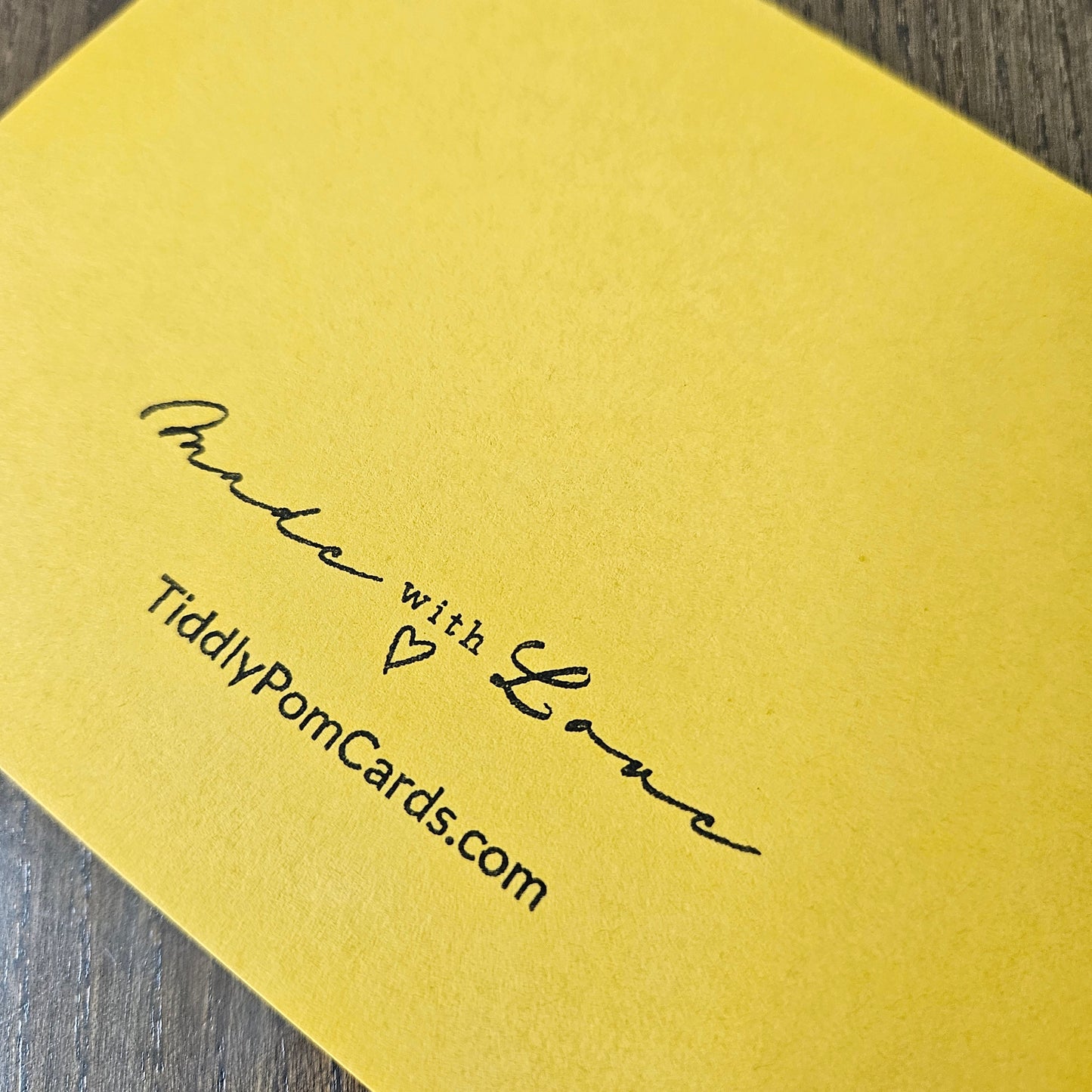 A close-up of a bright yellow greeting card showing the printed text 'Proud with Love' and 'TiddlyPomCards.com' in black ink, placed on a dark wood surface.