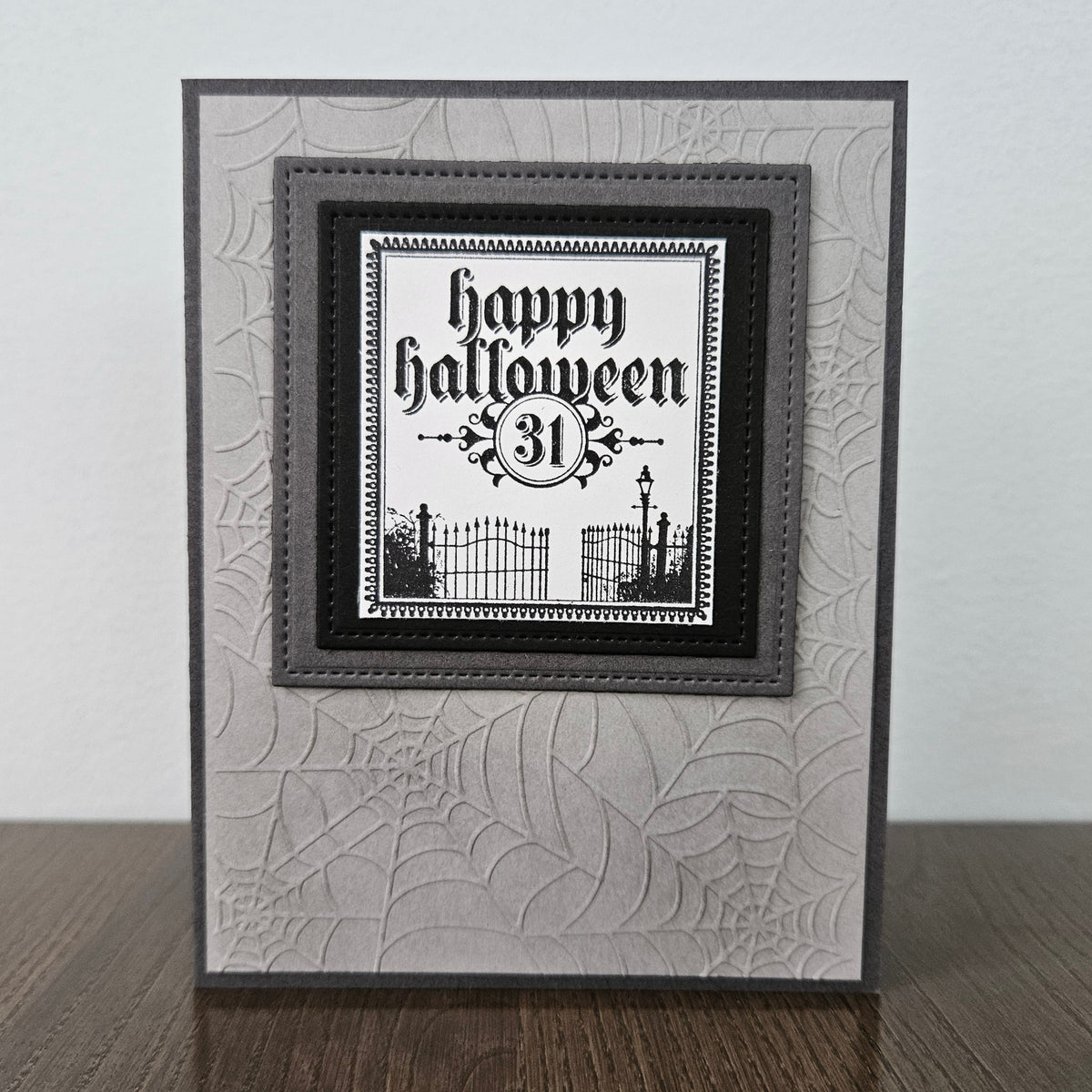 A handmade square greeting card with a Halloween theme, featuring a black and white 'Happy Halloween 31st' text design layered over a gray and white spiderweb-patterned card base, displayed on a wooden surface against a light gray wall.