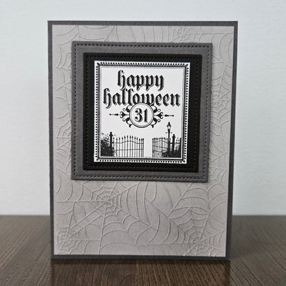 A handmade square greeting card with a Halloween theme, featuring a black and white 'Happy Halloween 31st' text design layered over a gray and white spiderweb-patterned card base, displayed on a wooden surface against a light gray wall.