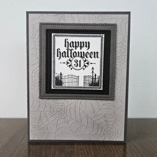 A handmade square greeting card with a Halloween theme, featuring a black and white 'Happy Halloween 31st' text design layered over a gray and white spiderweb-patterned card base, displayed on a wooden surface against a light gray wall.