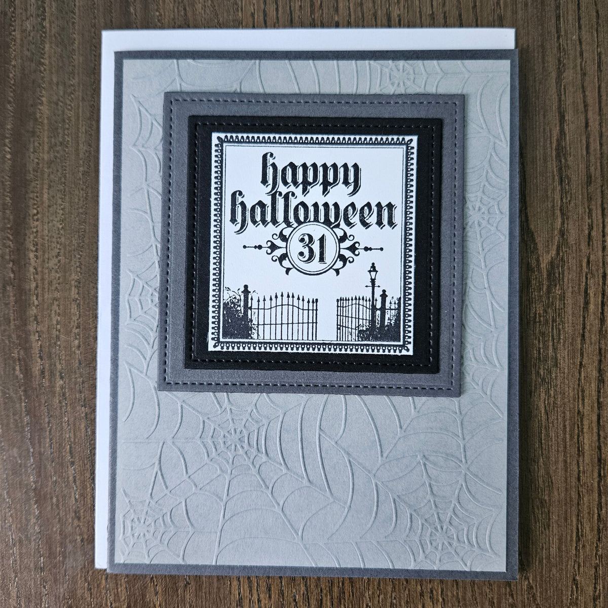A handmade square greeting card with a layered gray and black design, featuring an embossed spiderweb pattern on a light gray background. The center has a black and white label that reads 'happy halloween 31' with a spooky graveyard illustration.