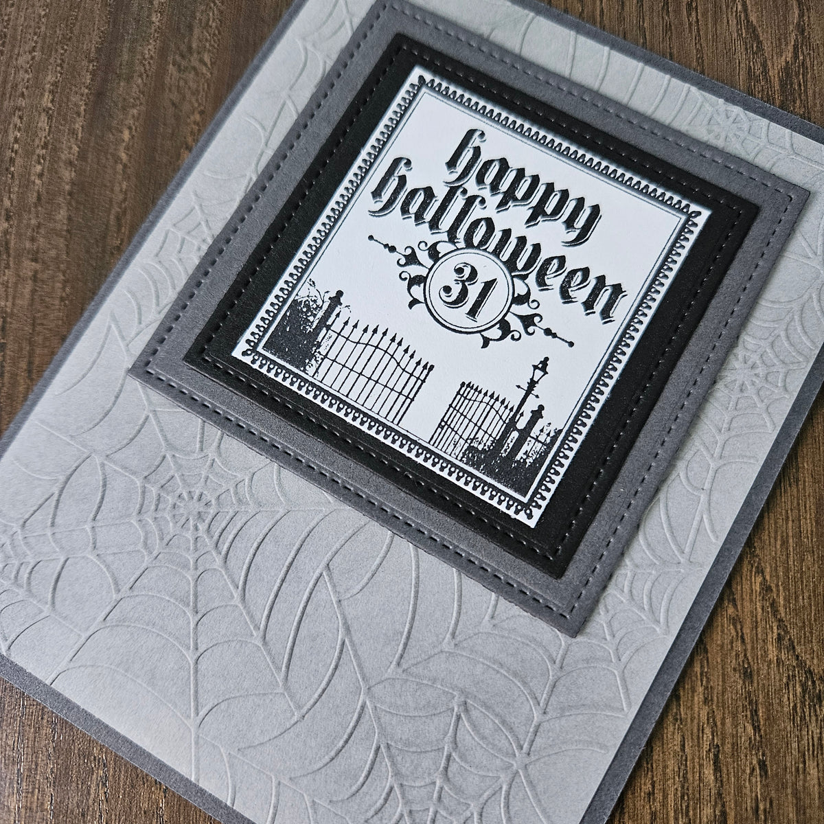 A close-up of a handmade Halloween greeting card featuring a layered black and white design with 'Happy Halloween 31' text, set on a wood surface.
