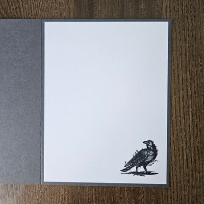 A square white art print or card with a dark gray border, featuring a black ink sketch of a raven near the bottom right corner. The print is displayed flat on a dark brown wood-grain surface, with a textured dark gray paper sheet partially visible on the left.