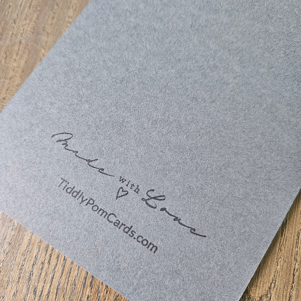 A close-up of a gray textured pom pom card on a wooden surface, printed with the brand name 'Tidily Pom Cards' and the phrase 'Made with Love'.