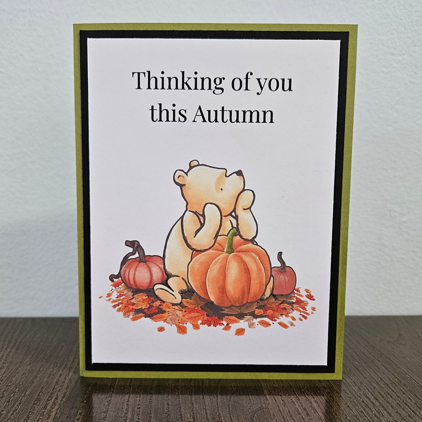 Front of a handmade A2 autumn card featuring classic Winnie the Pooh sitting with pumpkins and fall leaves framed by black and green borders with text reading Thinking of you this Autumn.