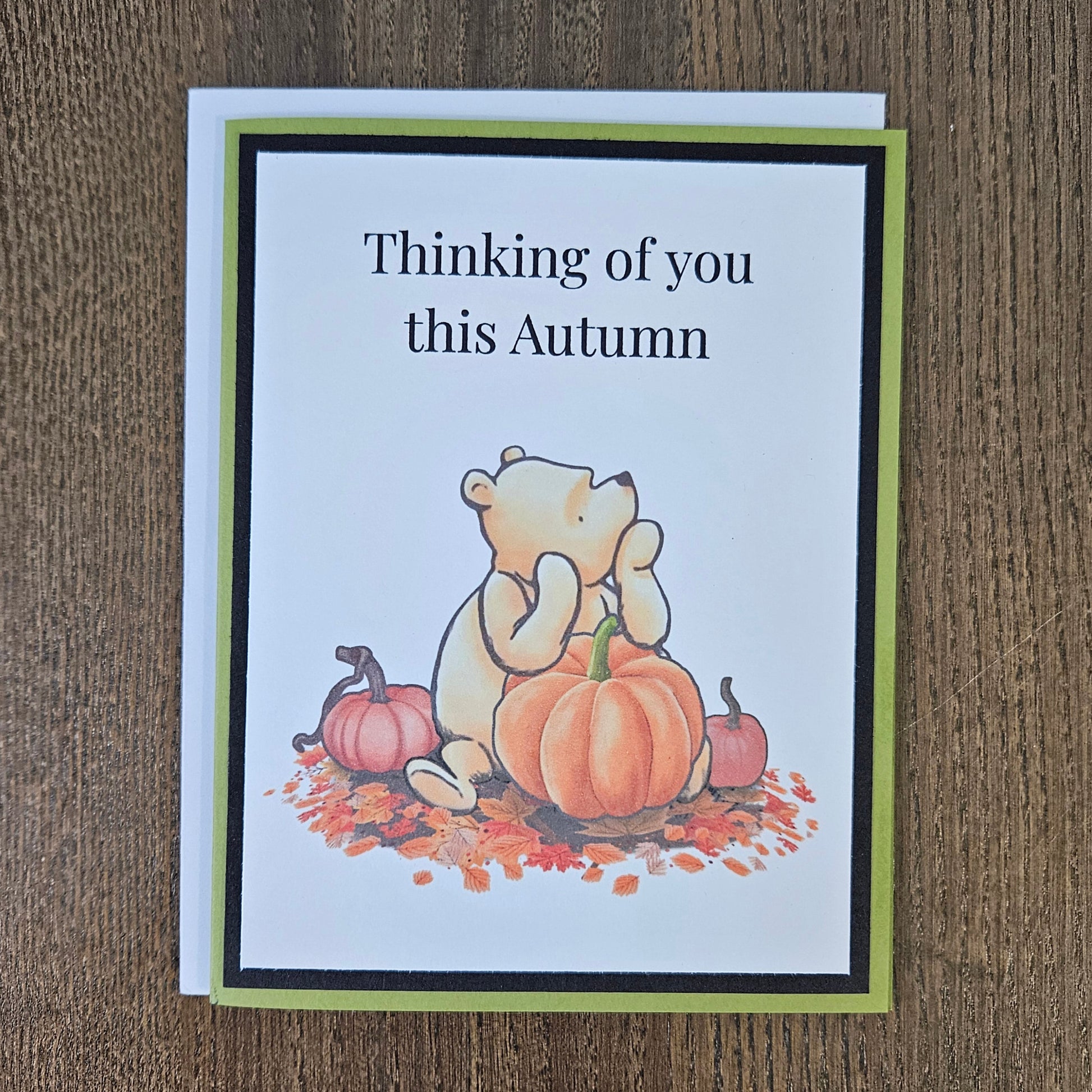 An autumn-themed greeting card featuring a cartoon bear sitting among pumpkins and fall leaves, with the text 'Thinking of you this Autumn' on a white card with a green and black border, placed on a wooden surface.