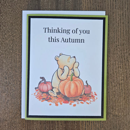 An autumn-themed greeting card featuring a cartoon bear sitting among pumpkins and fall leaves, with the text 'Thinking of you this Autumn' on a white card with a green and black border, placed on a wooden surface.