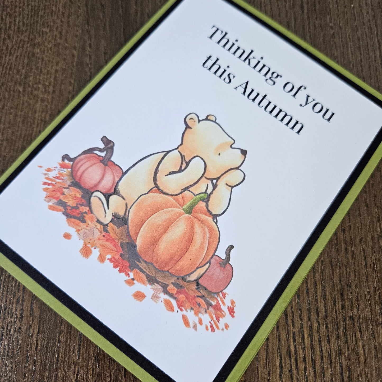 An autumn-themed greeting card featuring a teddy bear illustration with pumpkins and falling leaves, with the text 'Thinking of you this Autumn', displayed on a wooden surface.