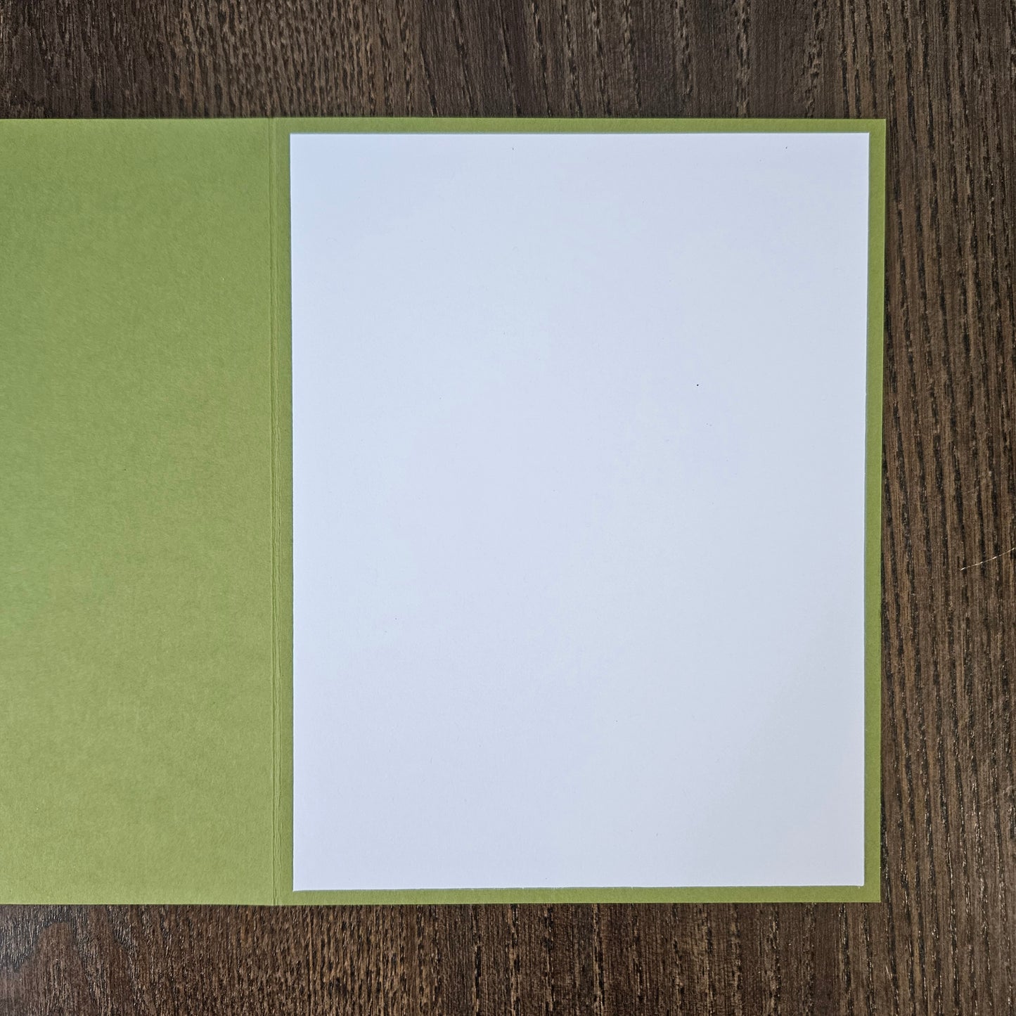 A blank white writing card or notepad page with a light olive green border, displayed open on a dark brown wood-grain surface.
