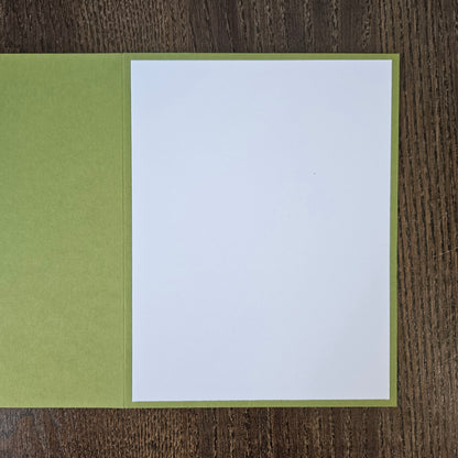 A blank white writing card or notepad page with a light olive green border, displayed open on a dark brown wood-grain surface.