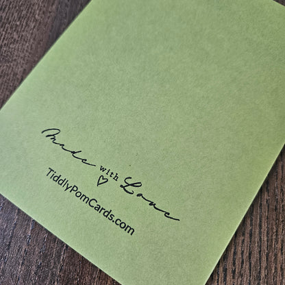 A square light green card with black printed text reading 'Pude with Love' and 'TidilyPornCards.com' placed on a dark wood surface.