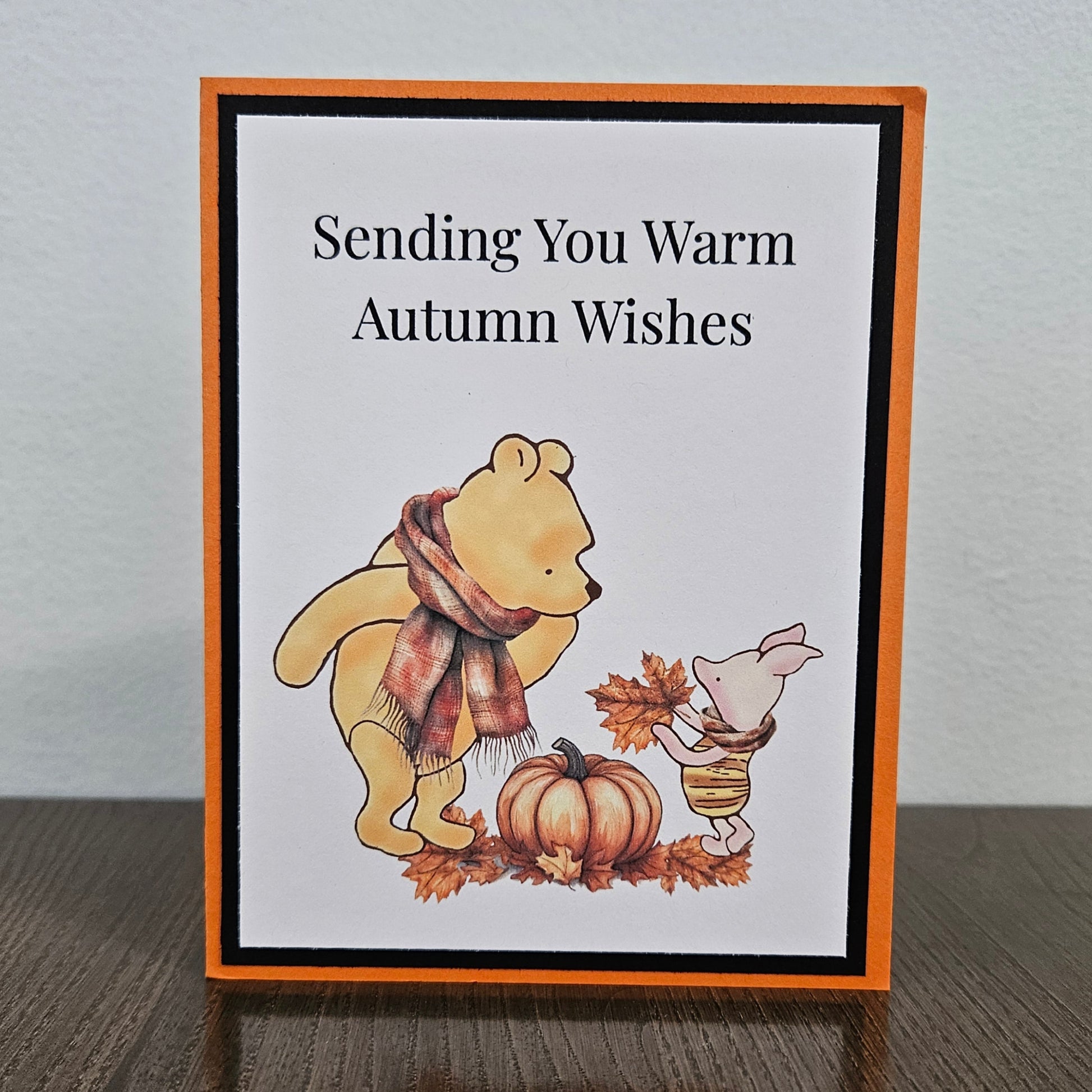 A handmade autumn-themed greeting card standing on a dark wooden table against a light gray wall. The card features a white background with the text 'Sending You Warm Autumn Wishes' in black, bordered by a black inner frame and an orange outer border.