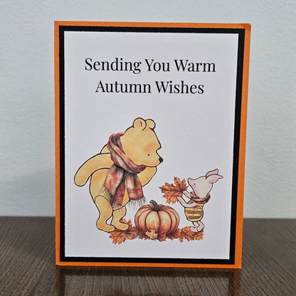 A handmade autumn-themed greeting card standing on a dark wooden table against a light gray wall. The card features a white background with the text 'Sending You Warm Autumn Wishes' in black, bordered by a black inner frame and an orange outer border.